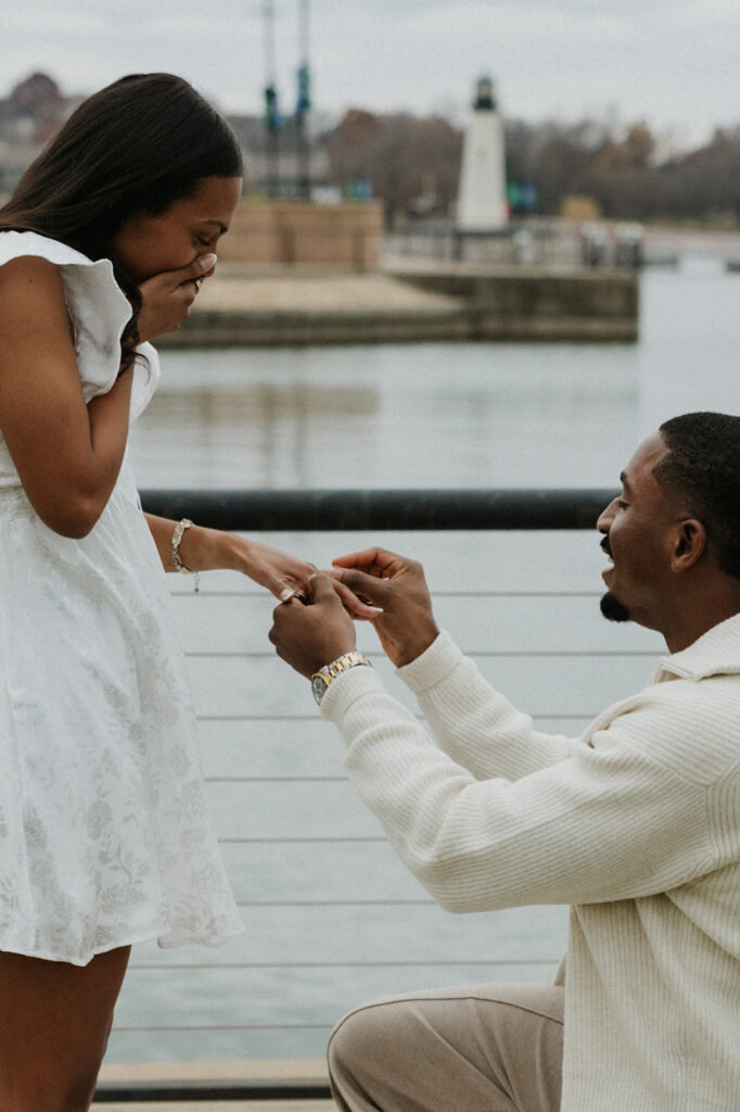 A man putting an engagement ring on his girlfriend's hand as she gasps in excitement at The Harbor in Rockwall.