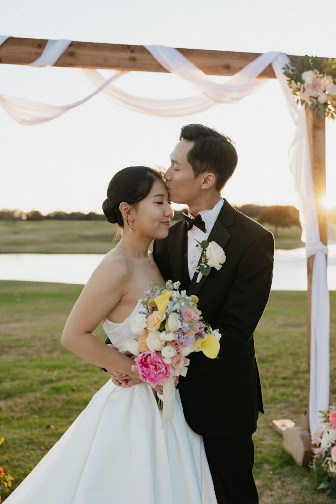 Groom kissing the bride's forehead as she holds a large, colorful bridal bouquet with pink and yellow flowers.