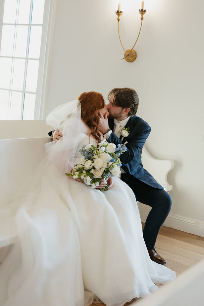 Bride and groom kissing romantically while sitting on a pew in the wedding chapel at The Nest at Ruth Farms.