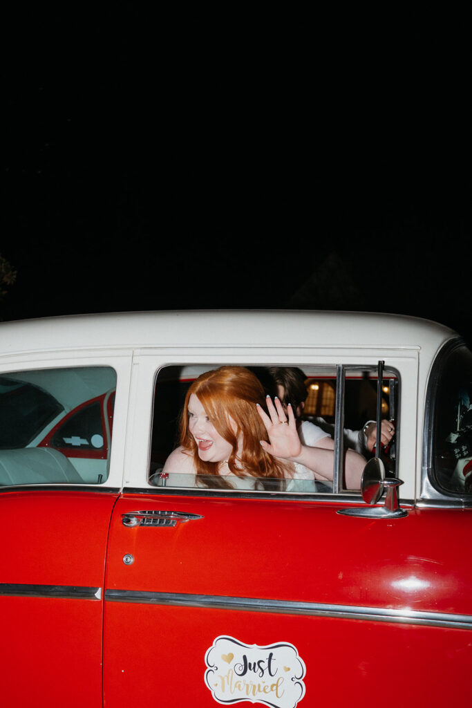 Bride waving goodbye to guests from a vintage car. 