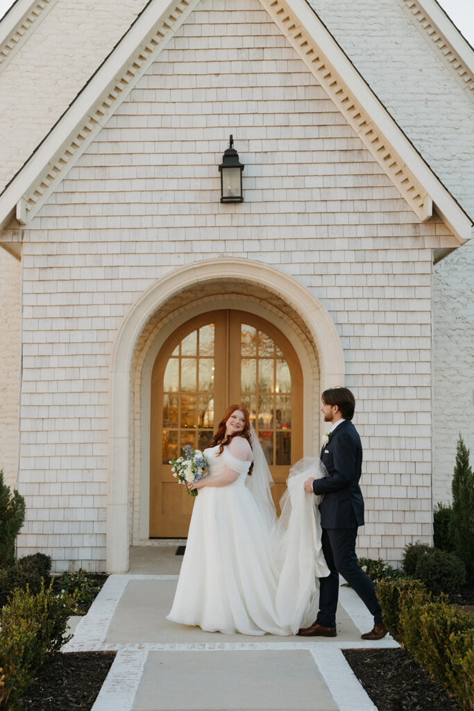 Groom holding his bride's train as they walk in front of the chapel at The Nest at Ruth Farms.