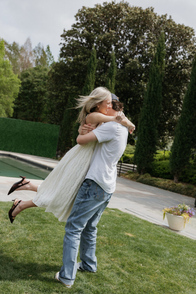 A woman with her arms wrapped around her fiancé as he spins her during their Dallas Arboretum and Botanical Garden proposal.