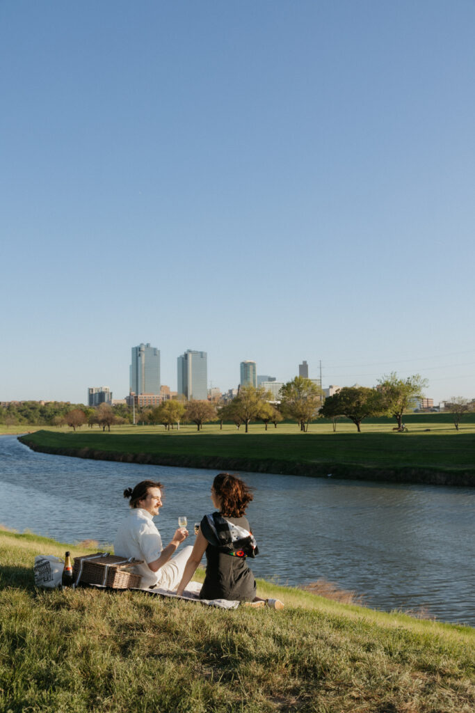 A couple having a picnic and drinking champagne by Trinity River, one of the best places to propose in Fort Worth.