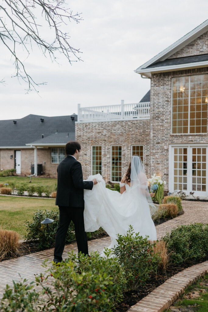 Groom holding his bride's train as they walk into their DFW wedding venue. 