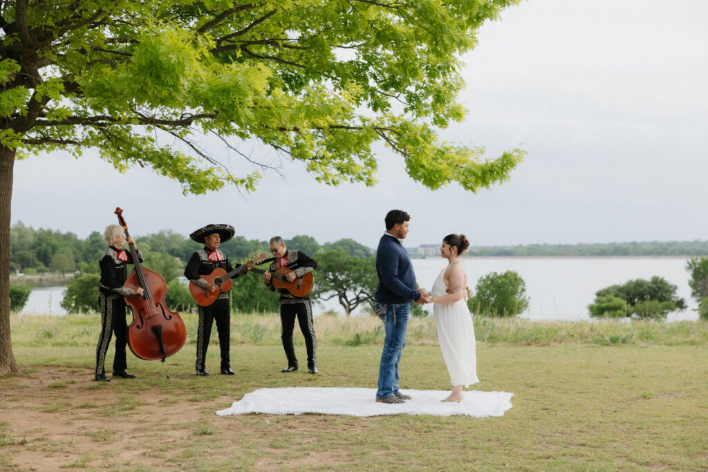 Couple holding hands while standing on a white blanket as a mariachi band plays.