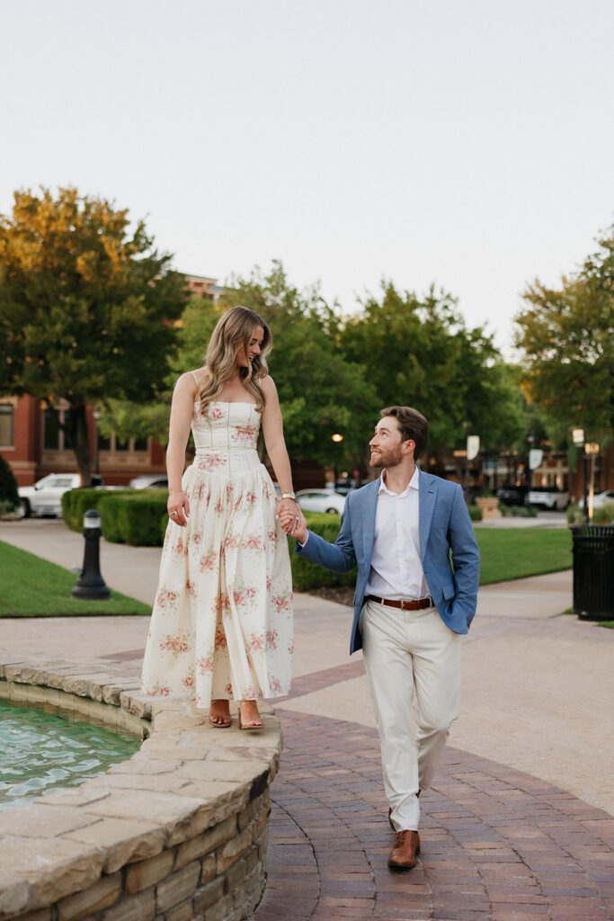 Woman standing on a fountain while her Fiancé holds her hand.